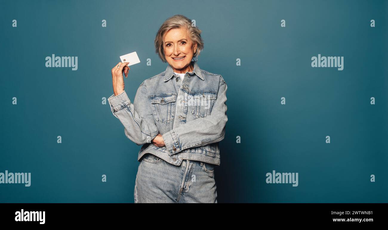 Femme âgée avec un sourire joyeux se tient gracieusement sur un fond bleu, tenant une carte de crédit dans sa main. Femme senior appréciant la finance moderne Banque D'Images