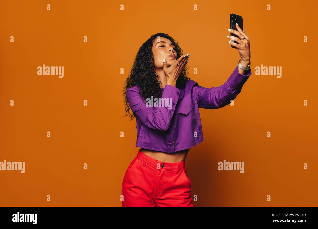 Femme à la mode, avec des cheveux bouclés et des vêtements orange vif, se tient sur un fond vibrant. Tenant un téléphone portable, elle pose en toute confiance et Banque D'Images