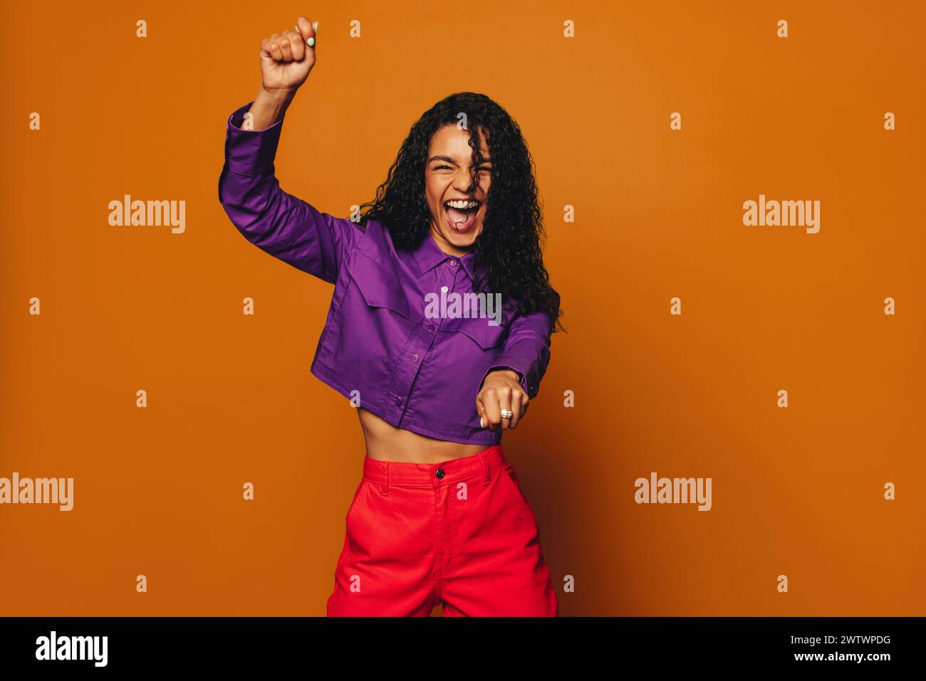 Femme joyeuse avec les cheveux bouclés, vêtements décontractés élégants se tient sur fond orange vibrant. Célébrant et s'amusant, elle danse joyeusement, rad Banque D'Images