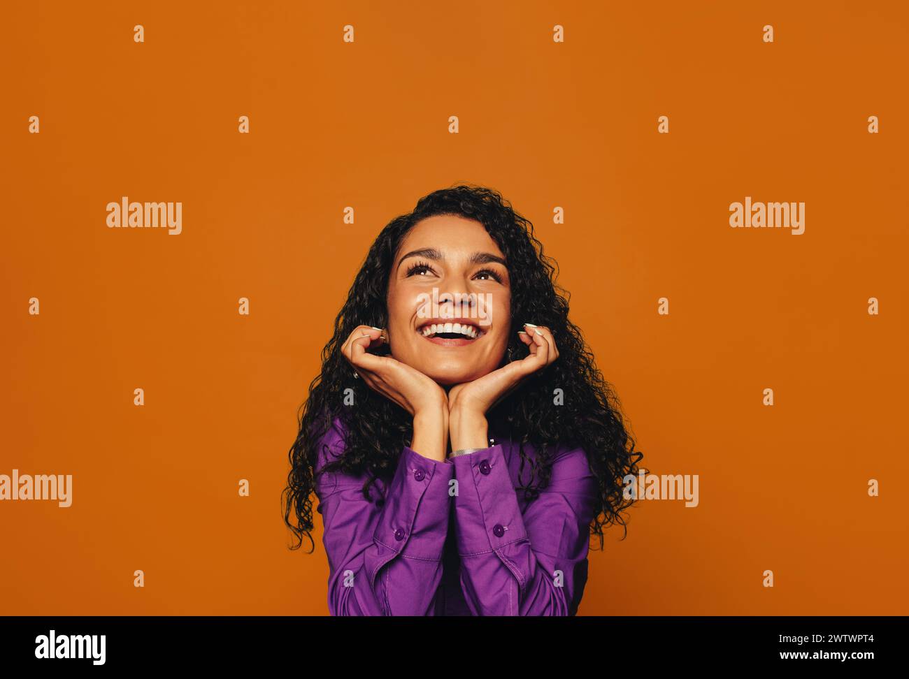 Femme heureuse aux cheveux bouclés, portant des vêtements décontractés, souriant avec une expression rêveuse. Sur fond orange, son expression pensive mais heureuse i Banque D'Images