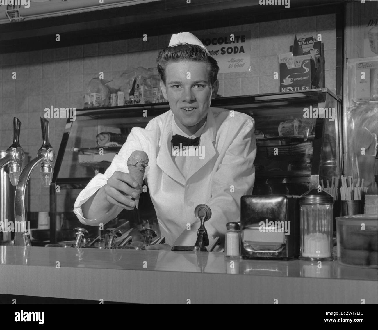 Jeune homme vêtu d'un manteau blanc, d'un noeud papillon et d'un chapeau blanc, souriant à la caméra tout en se tenant debout derrière le comptoir d'un magasin de boissons gazeuses offrant un cornet de crème glacée et un sourire Banque D'Images