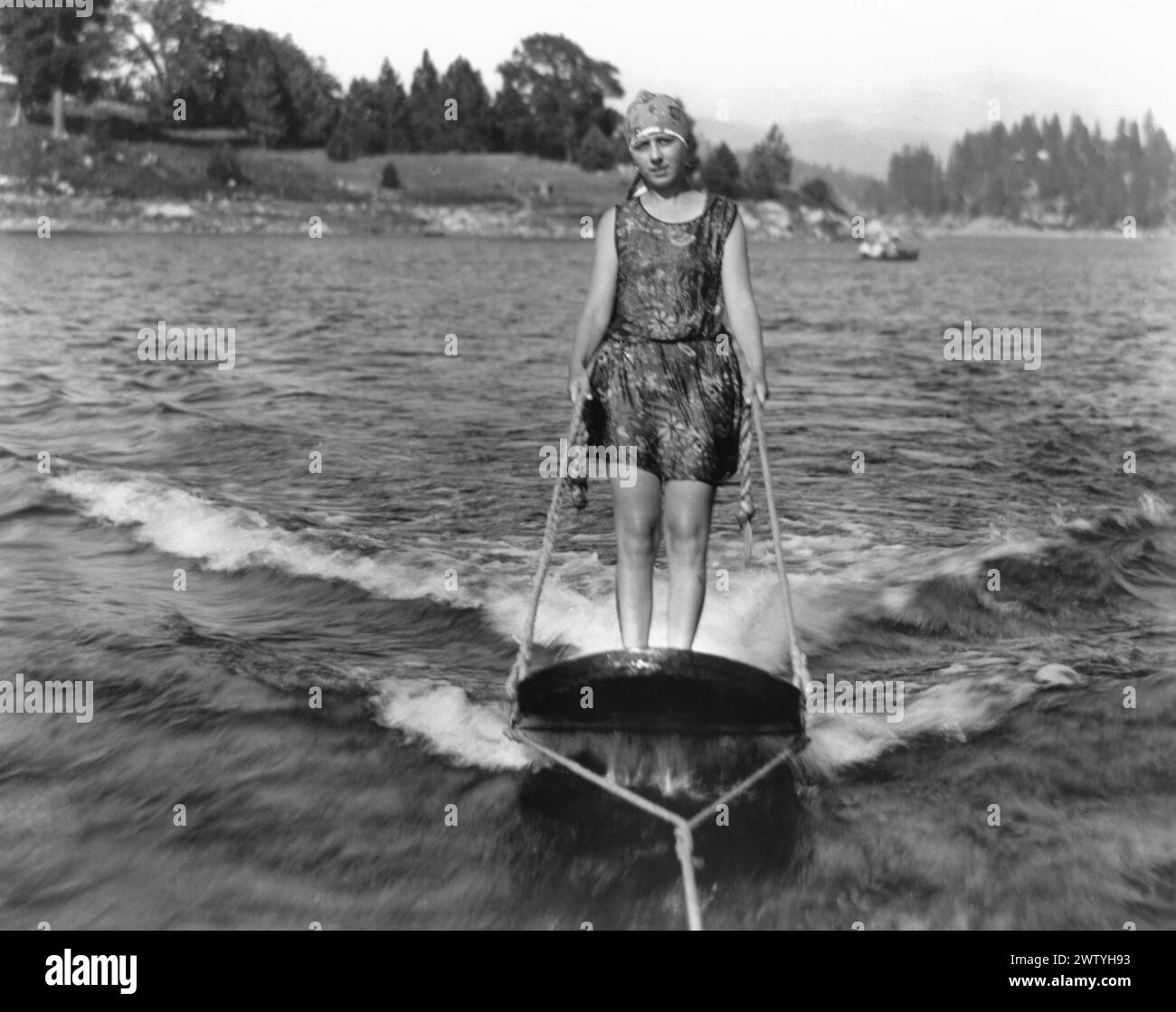 femme des années 1920 en maillot de bain et casquette chevauchant sur planche de surf derrière un bateau Banque D'Images