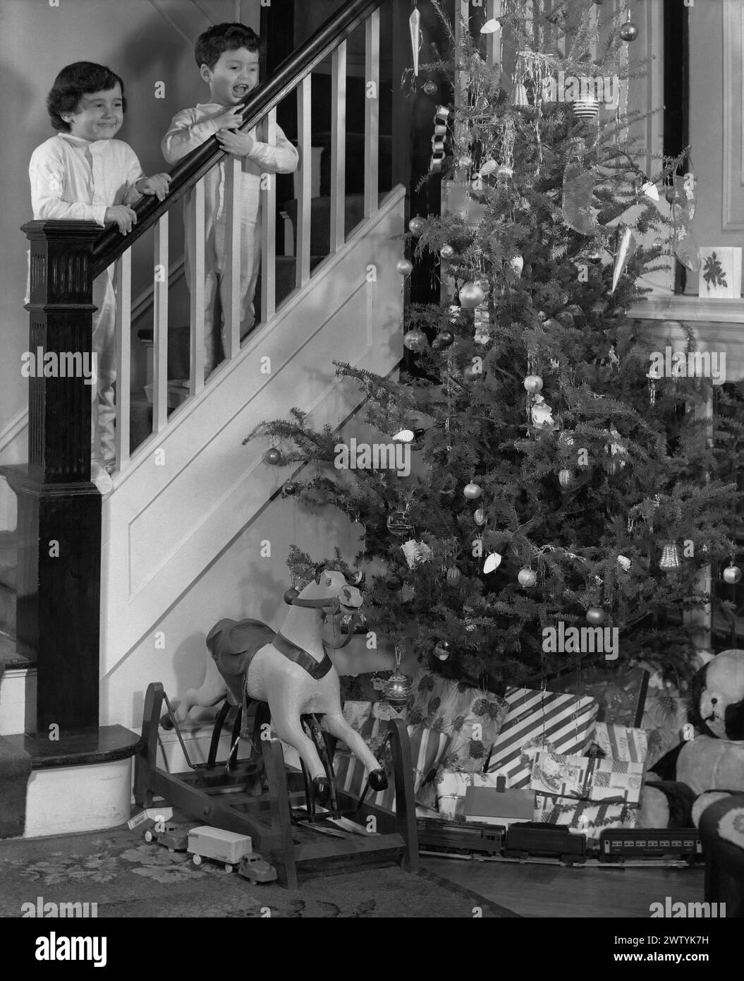 Jeune garçon et sa sœur en pyjama debout sur l'escalier à la rampe regardant vers le bas sur un arbre de Noël chargé de jouets et de cadeaux le matin de Noël Banque D'Images