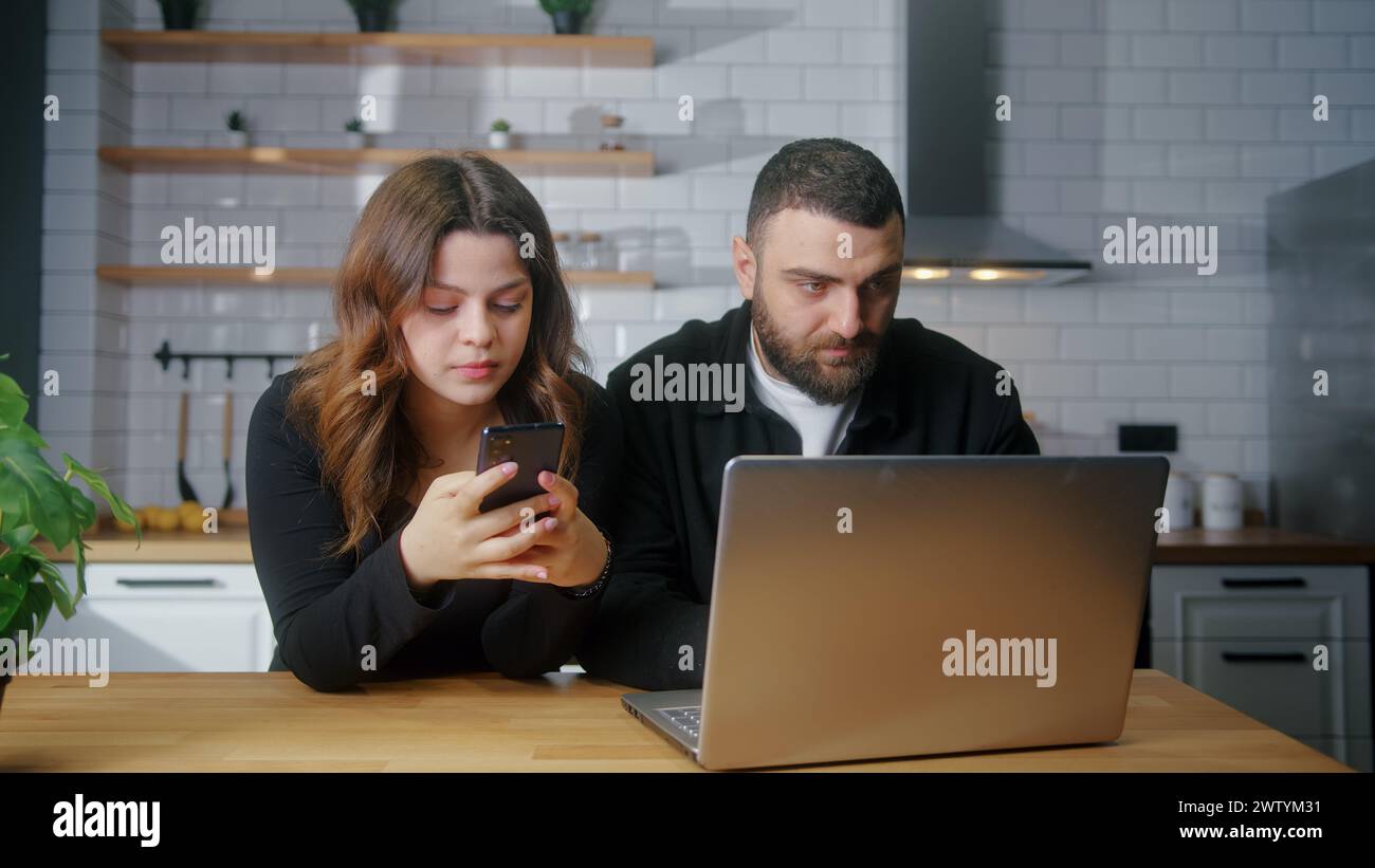 Jeune couple assis dans la cuisine femme à l'aide d'un smartphone, homme tapant à l'aide d'un ordinateur portable, passant du temps sur Internet à la maison Banque D'Images