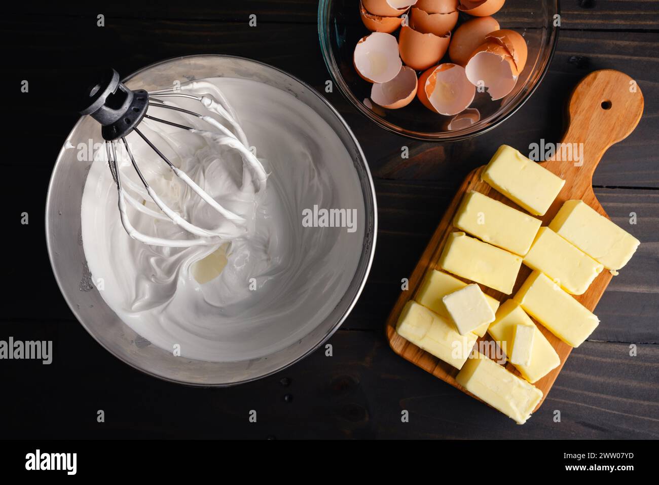 Bol à mélanger rempli de meringue à côté de bâtonnets de beurre : fouet métallique placé dans un bol plein de meringue suisse près des œufs cassés et du beurre Banque D'Images