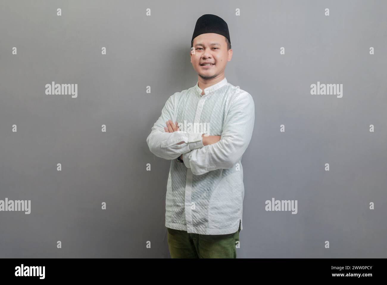 Homme musulman asiatique debout avec le bras croisé, portant une chemise koko et peci avec des nuances du mois de jeûne, isolé sur un fond gris Banque D'Images