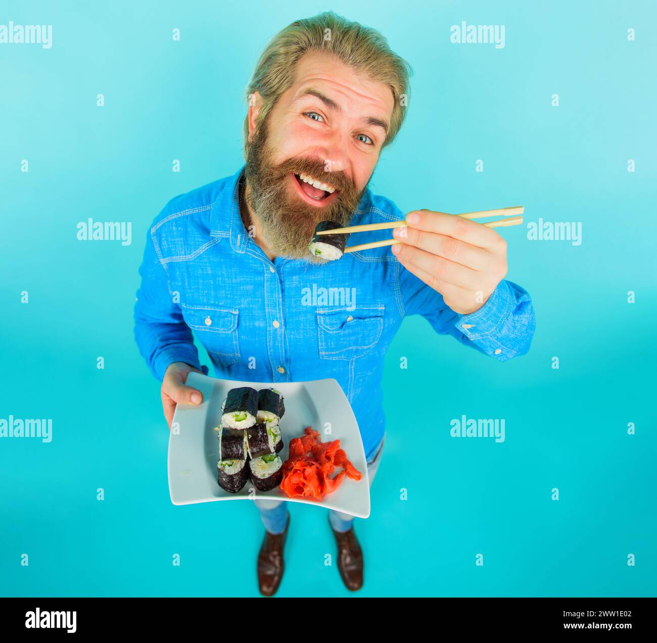 Livraison de sushis. Cuisine asiatique. Homme barbu souriant mangeant un savoureux rouleau de sushi avec des baguettes. Cuisine japonaise. Beau homme en chemise en denim avec plaque Banque D'Images