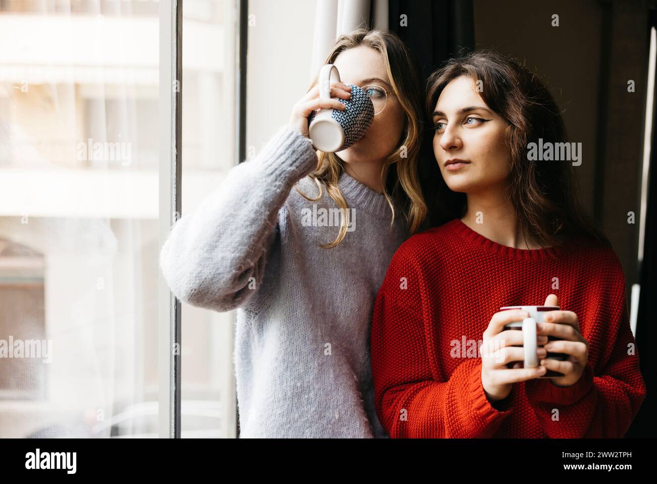 Deux jeunes femmes debout côte à côte, tenant une tasse de thé, près d'une fenêtre à l'intérieur de leur maison, partageant un moment de compagnie dans un et intime Banque D'Images