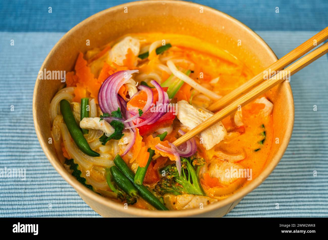 Curry rouge épicé avec légumes, viande de poulet et lait de cacao dans un bol et baguettes sur fond bleu, gros plan. Cuisine asiatique. Banque D'Images