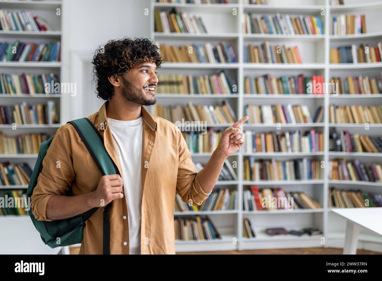 Un jeune adulte dans une tenue décontractée avec un sac à dos pointant vers quelque chose dans une bibliothèque pleine de livres. Banque D'Images