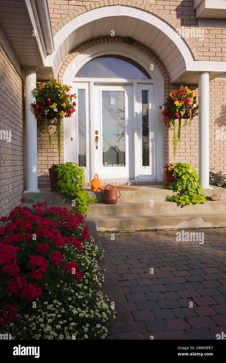 Porte d'entrée avant sur brique tan avec portique de garniture blanche et bordure surélevée avec pélargonium rouge, fleurs de géranium en été, Québec, Canada Banque D'Images
