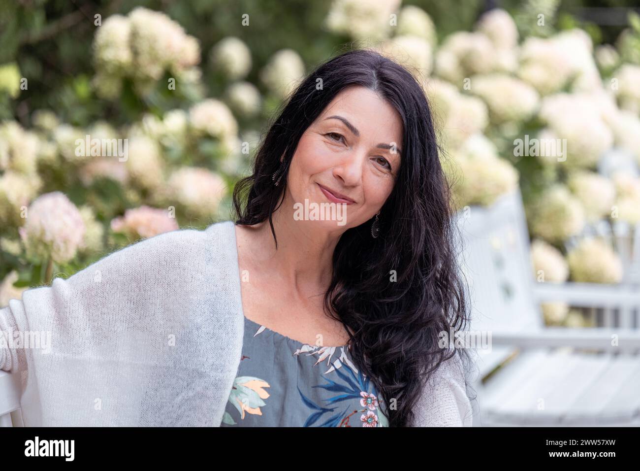 Une femme souriante aux cheveux noirs, portant une robe florale et un châle blanc, est assise sur un banc de parc entouré Banque D'Images