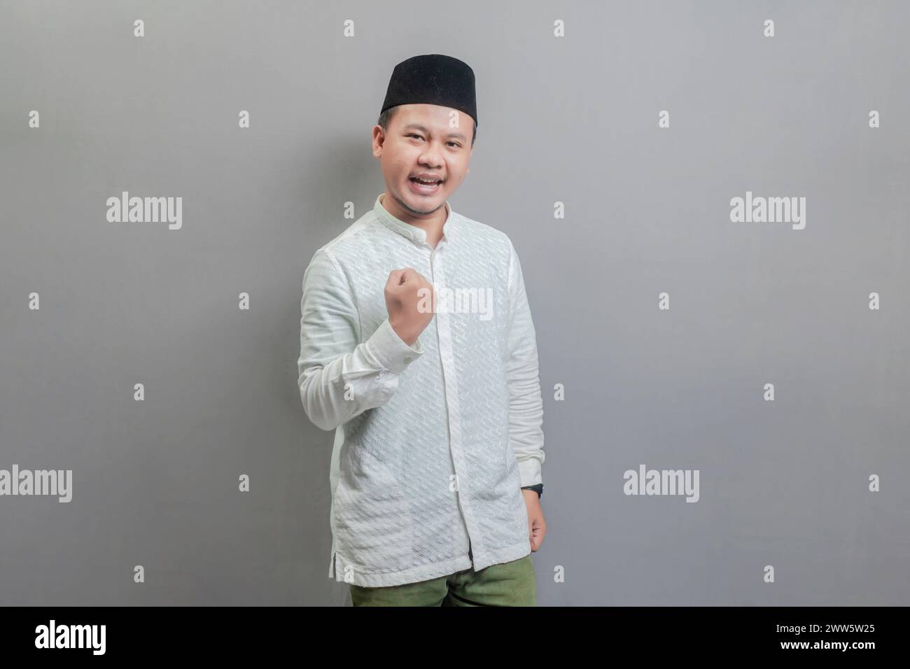Heureux homme musulman asiatique amusant réussi main levée portant une chemise koko et peci avec des nuances du mois de jeûne, isolé sur un fond gris Banque D'Images