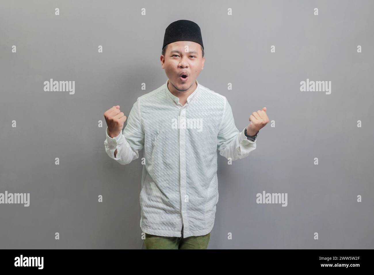 Heureux homme musulman asiatique amusant réussi main levée portant une chemise koko et peci avec des nuances du mois de jeûne, isolé sur un fond gris Banque D'Images