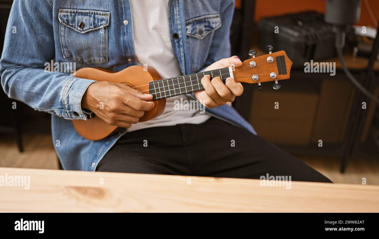 Homme hispanique jouant du ukulélé à l'intérieur, portant du denim, assis par une table en bois dans un cadre de studio décontracté. Banque D'Images