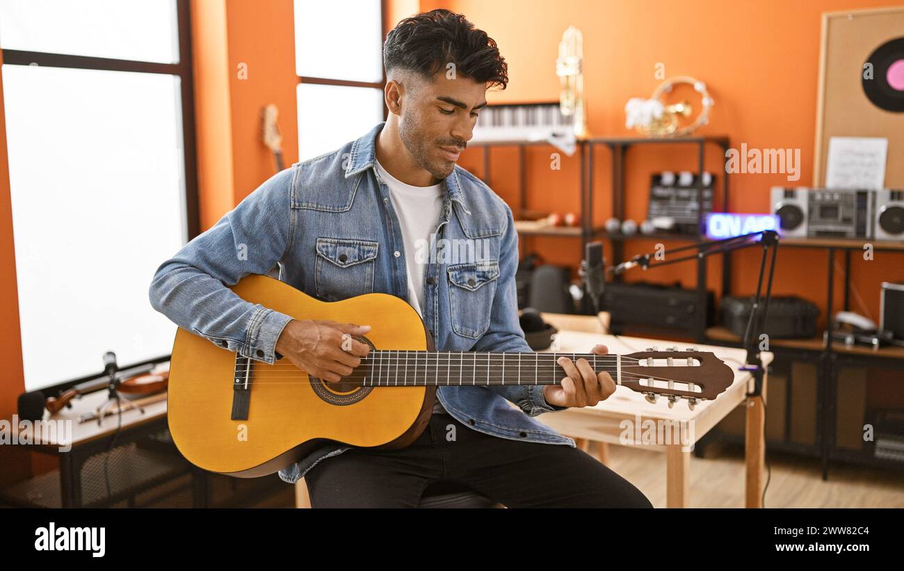 Bel homme hispanique barbu jouant de la guitare acoustique dans un studio d'enregistrement confortable avec microphone et équipement sonore. Banque D'Images