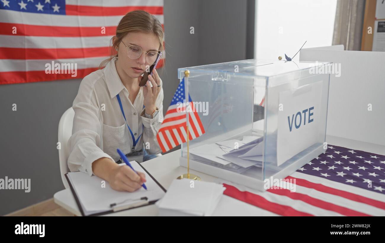 Une jeune femme caucasienne effectue plusieurs tâches, écrivant et parlant au téléphone dans un centre de vote électoral américain avec des drapeaux Banque D'Images