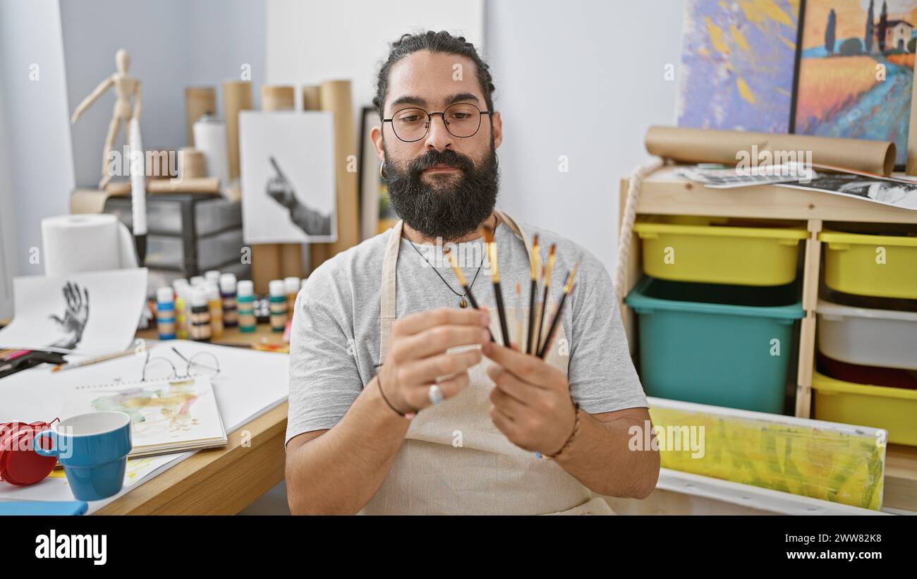 Un homme barbu examine des pinceaux dans un atelier d'art entouré de peintures, de toiles et de fournitures d'art. Banque D'Images