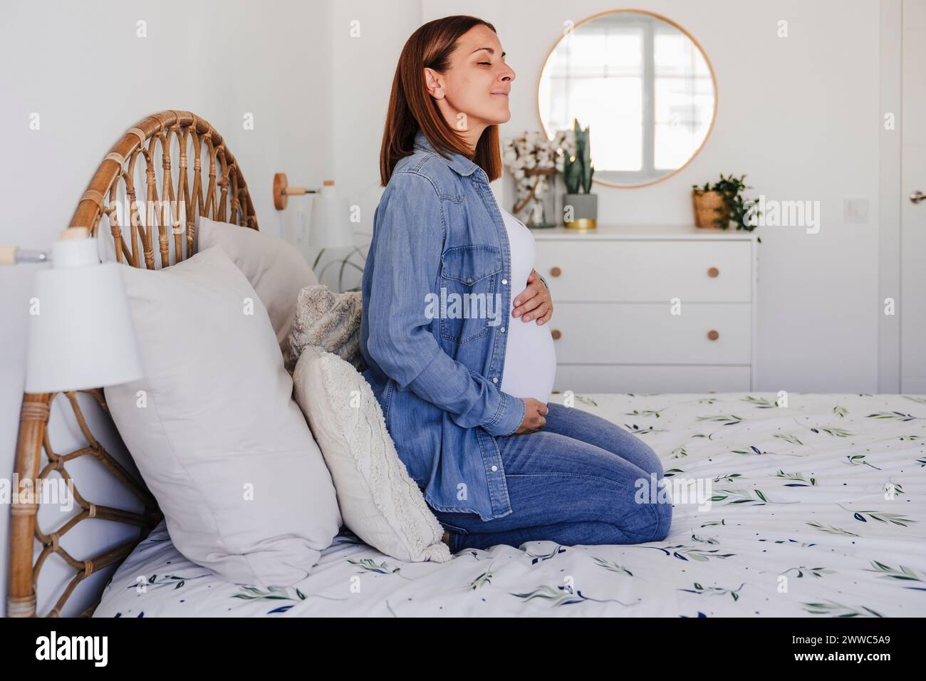 Femme enceinte avec les yeux fermés assis sur le lit à la maison Banque D'Images
