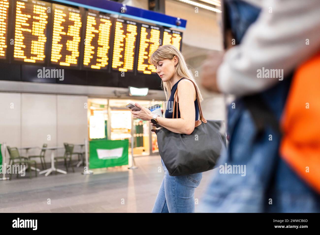 Femme utilisant un téléphone intelligent debout près du tableau de départ d'arrivée à la gare Banque D'Images