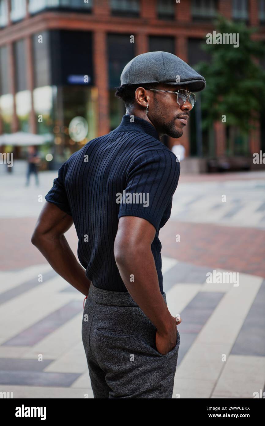 Bel homme afro-américain élégant se tient dans la rue dans une tenue à la mode, portant une casquette, des lunettes de soleil et un t-shirt noir, la mode masculine moderne Banque D'Images