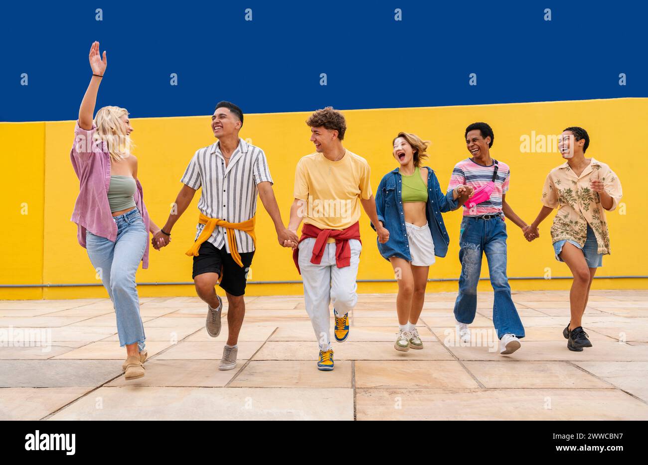 Groupe multiethnique de jeunes amis courant devant le mur jaune se tenant par la main Banque D'Images