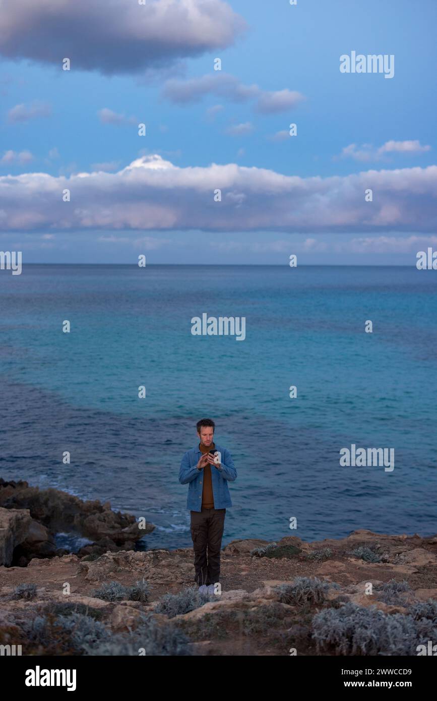Homme utilisant un téléphone intelligent debout sur la colline près de la mer au crépuscule Banque D'Images