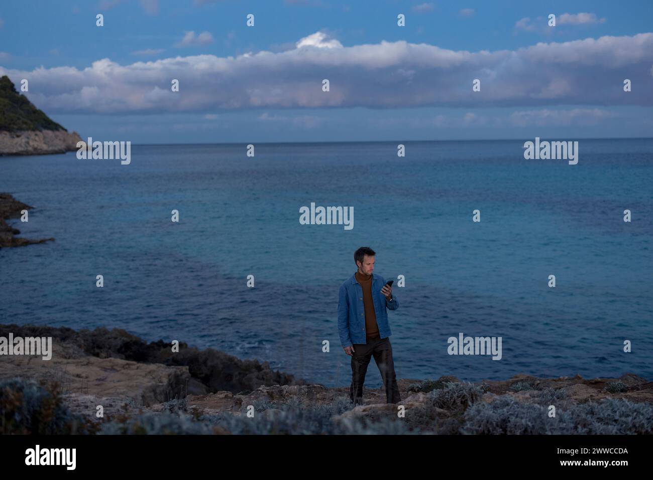 Homme utilisant un téléphone intelligent debout sur la colline près de l'océan au crépuscule Banque D'Images
