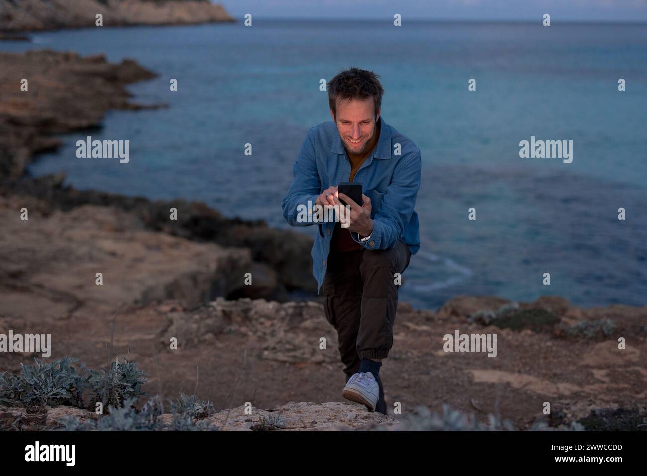 Homme souriant à l'aide d'un téléphone intelligent sur la colline près de la mer au crépuscule Banque D'Images