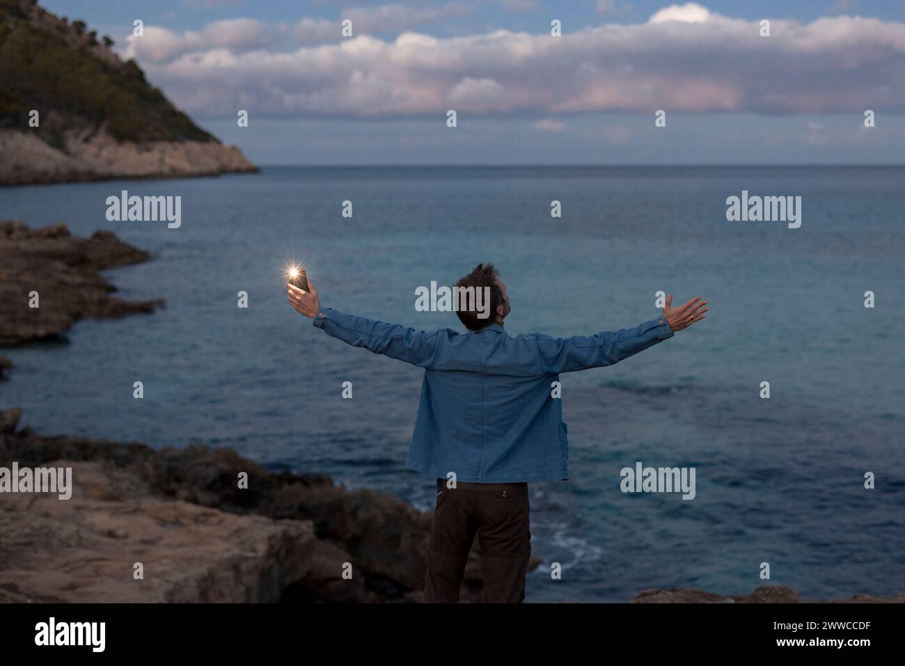 Homme insouciant avec les bras tendus debout sur la colline près de la mer au crépuscule Banque D'Images