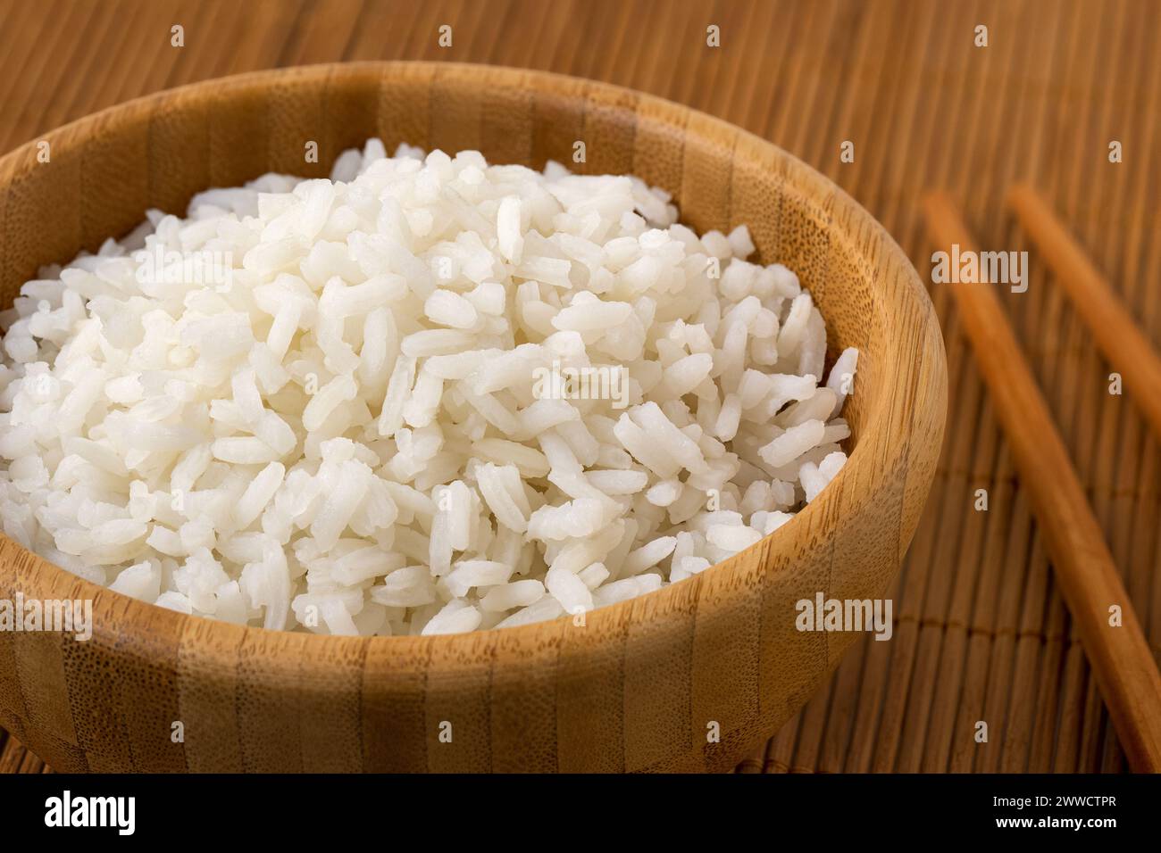 Détail de riz blanc cuit dans un bol en bois foncé à côté de baguettes sur bambou mat. Banque D'Images