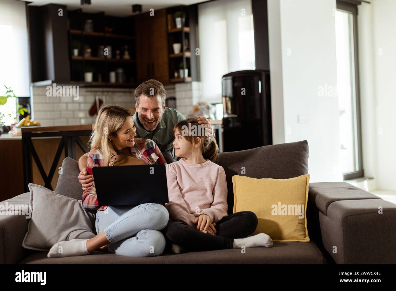 Joyeuse famille de trois passe du temps de qualité ensemble sur le canapé du salon, partageant un moment autour d'un ordinateur portable dans leur maison confortable Banque D'Images