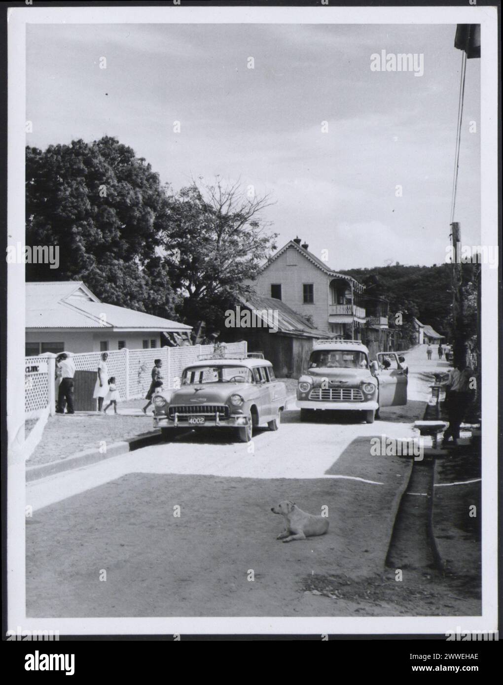 Description : Honduras britannique. [Scène de rue à Benque Viejo, del Carmen Cayo District. L'image montre deux voitures. L'un conduit vers la caméra tandis que l'autre est garé avec une famille marchant vers elle. Un chien est allongé sur la route]. Photographie n° R 34997 photographie officielle du Honduras britannique compilée par le Bureau central de l'information. Crown Copyright réservé. Lieu : Honduras britannique date : 1965 avr belize, caraïbes, caribbeanthrough alens Banque D'Images