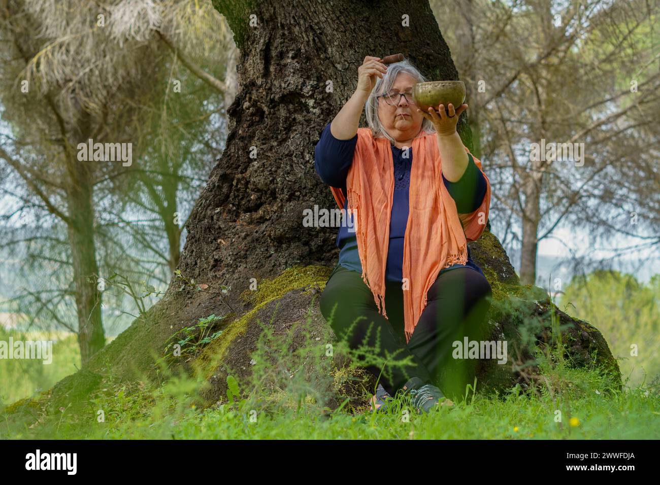 Femme mature aux cheveux blancs méditant dans la forêt avec les yeux fermés et un bol chantant tibétain qui sonne Banque D'Images
