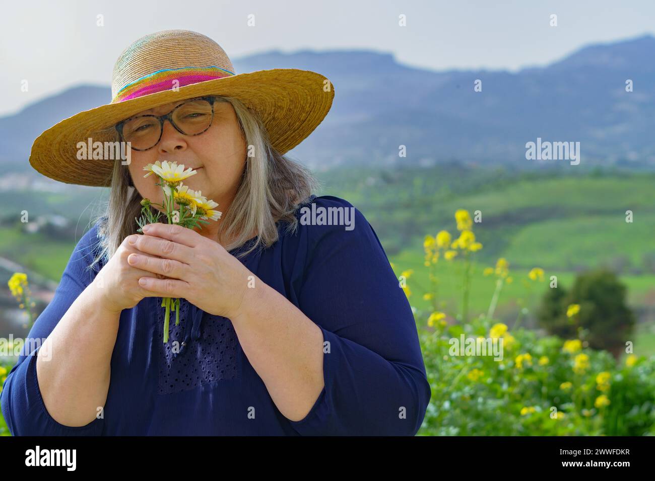 Femme mature avec les cheveux blancs et chapeau tenant un bouquet de marguerites dans ses mains dans le fond un paysage de montagne Banque D'Images