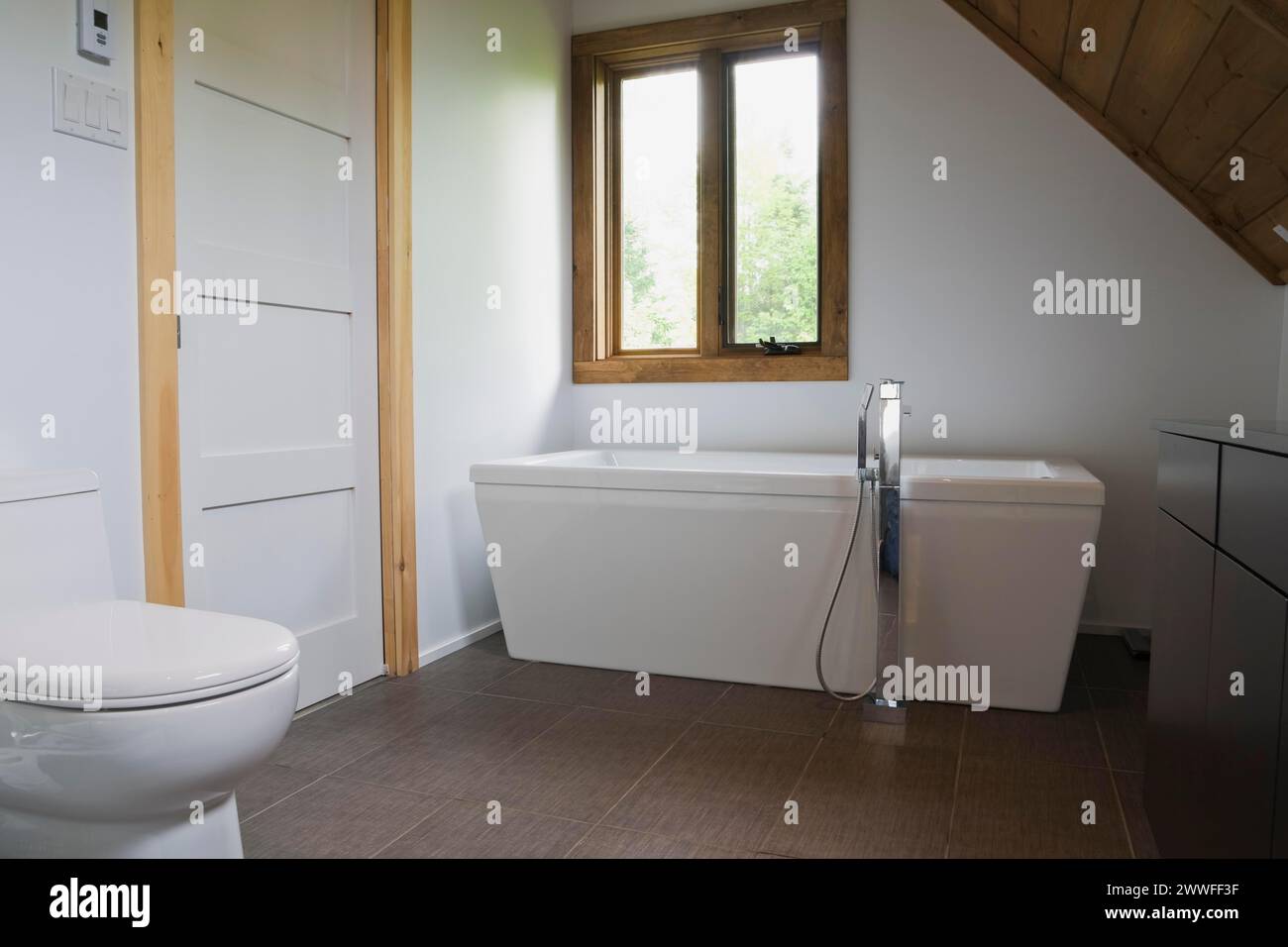 Salle de bains principale avec carrelage en céramique grise et toilettes en porcelaine blanche et baignoire autoportante à l'étage à l'intérieur de la maison en rondins de style contemporain Banque D'Images