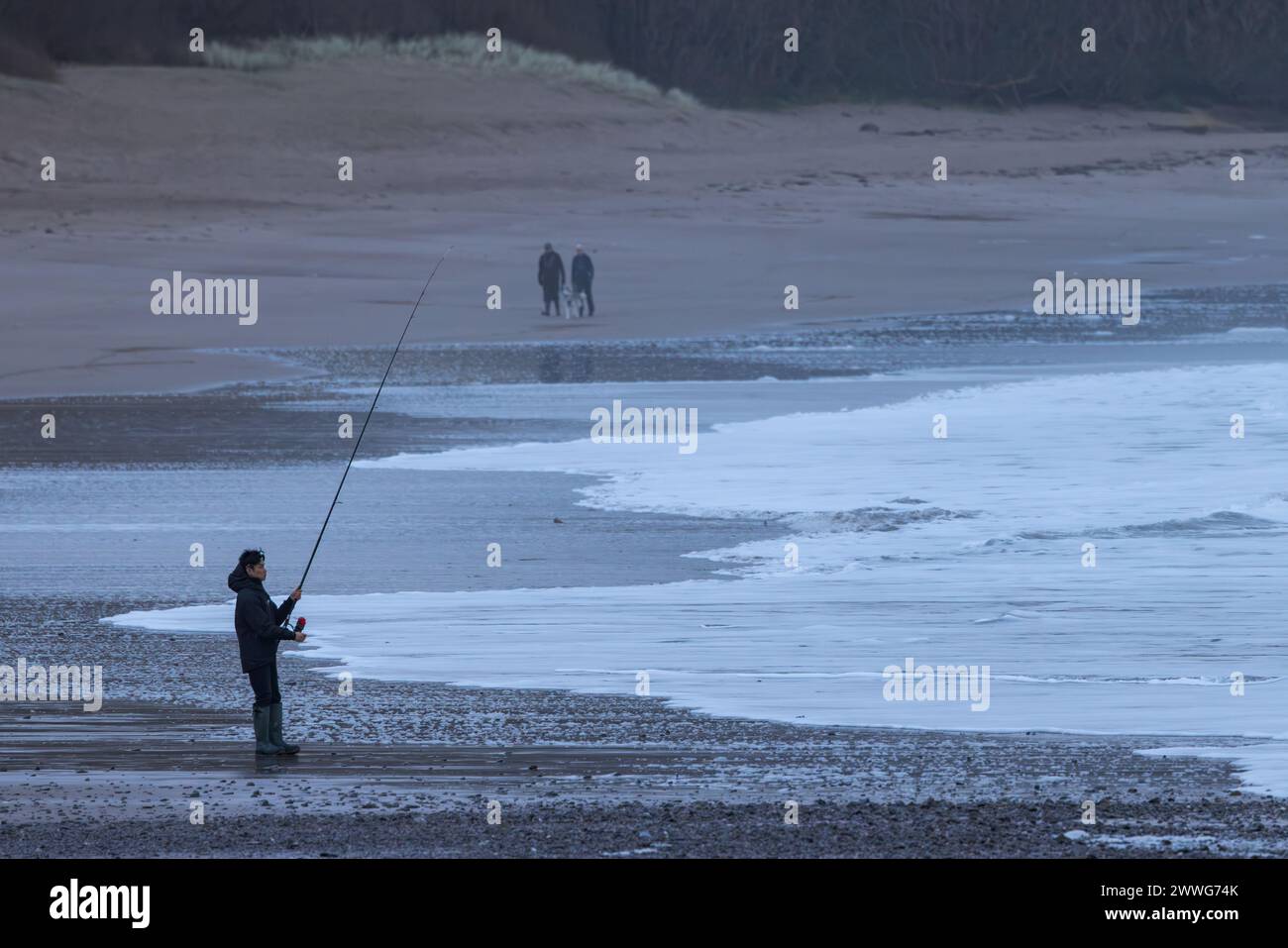 Homme pêchant de la plage est d'eau douce dans le Pembrokeshire, pays de Galles avec promeneurs de chiens en arrière-plan Banque D'Images