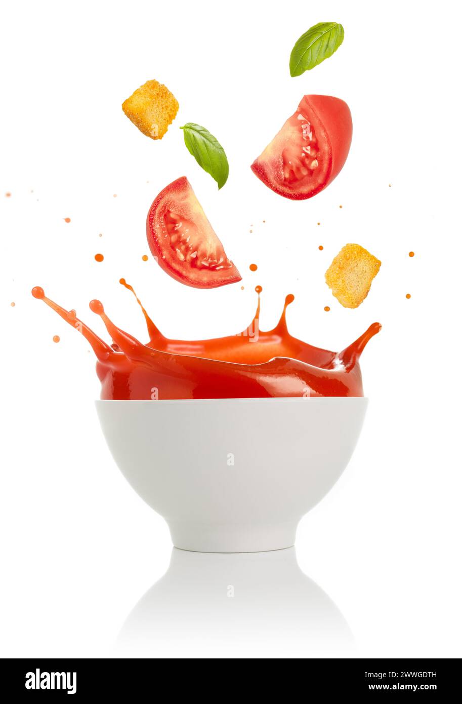 Tomates, croûtons et feuilles de basilic tombant dans un bol de soupe isolé sur fond blanc. Banque D'Images