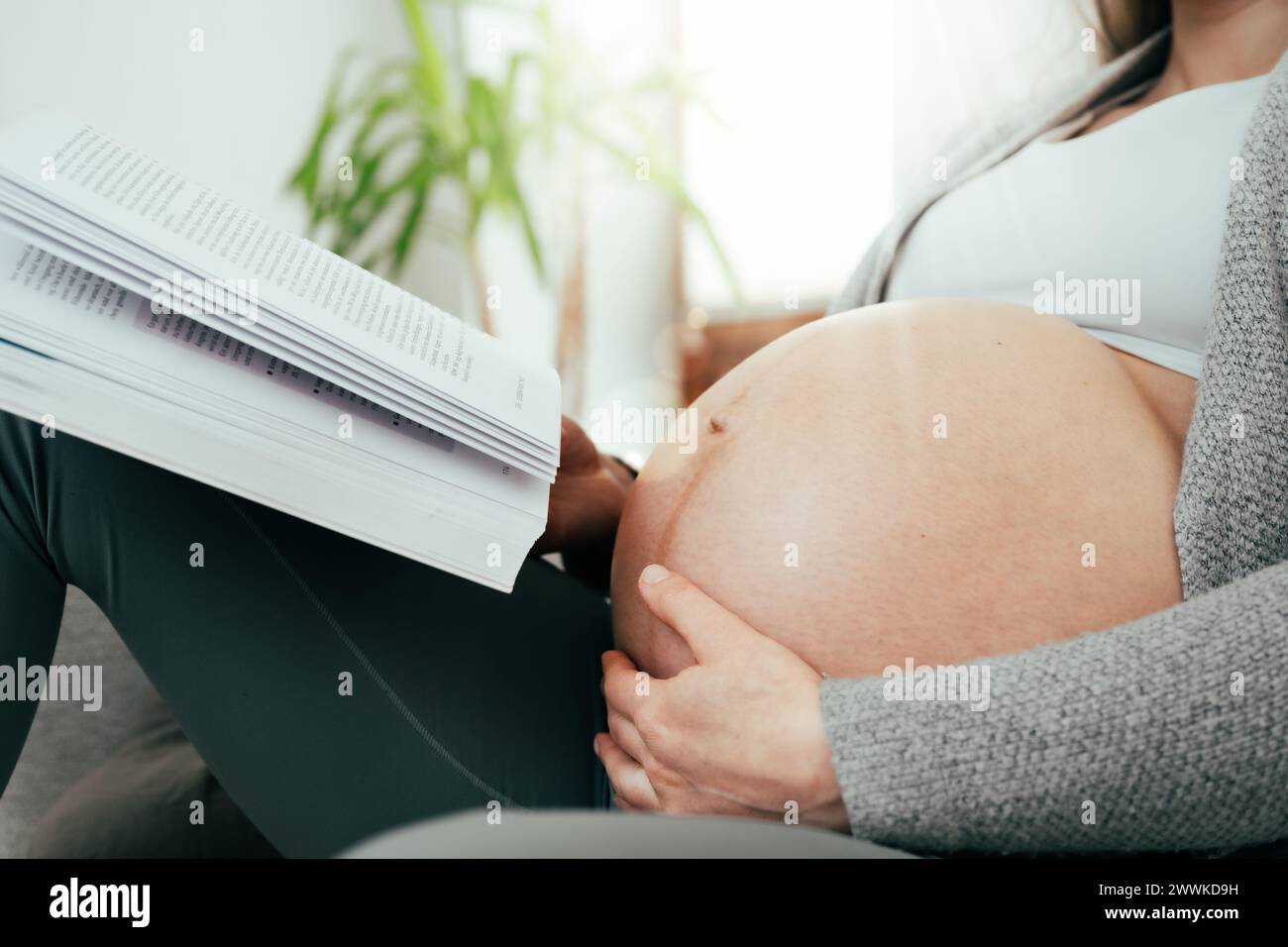 Description : angle bas de la femme dans les derniers mois de grossesse tenant doucement son ventre tout en lisant un livre. Grossesse troisième trimestre - semaine 34 Banque D'Images