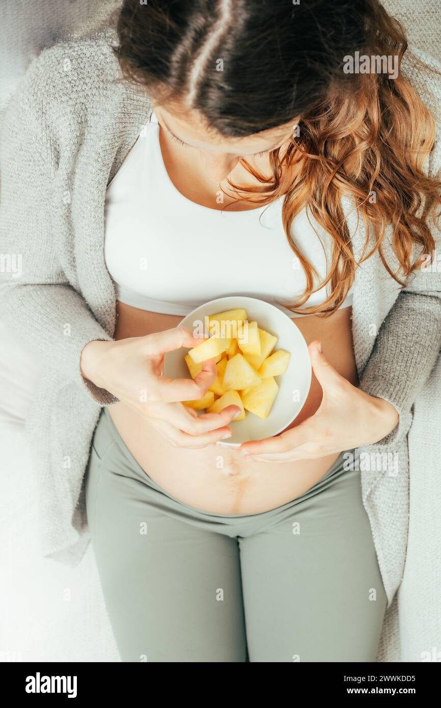 Description : vue de dessus d'une femme assise sur le canapé mangeant des morceaux d'ananas tout en tenant doucement son ventre dans les derniers mois de grossesse. Grossesse th Banque D'Images