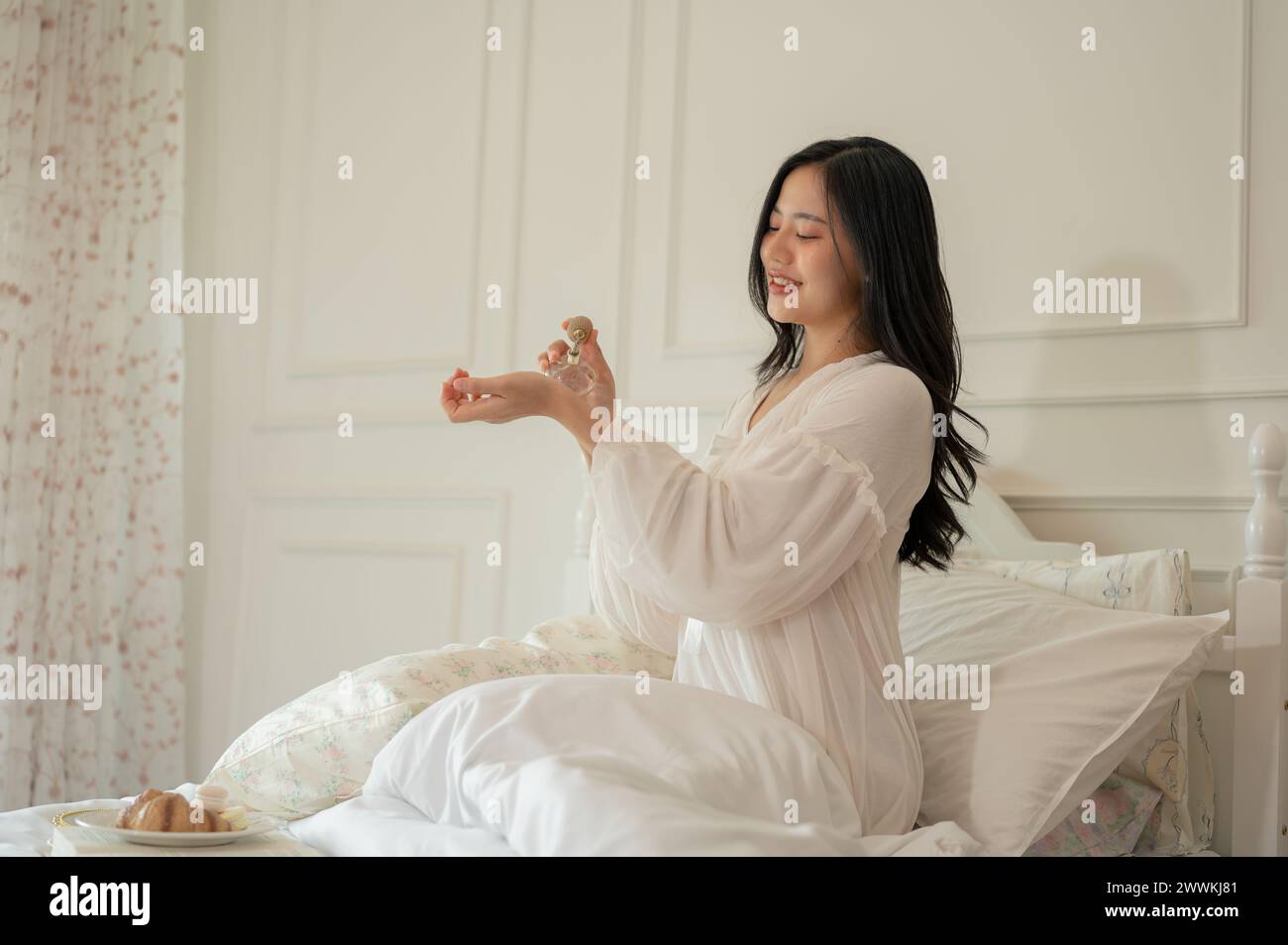 Une femme asiatique attrayante en pyjama à robe longue applique un parfum après s'être réveillée sur son lit. vie domestique et concepts de mode de vie Banque D'Images