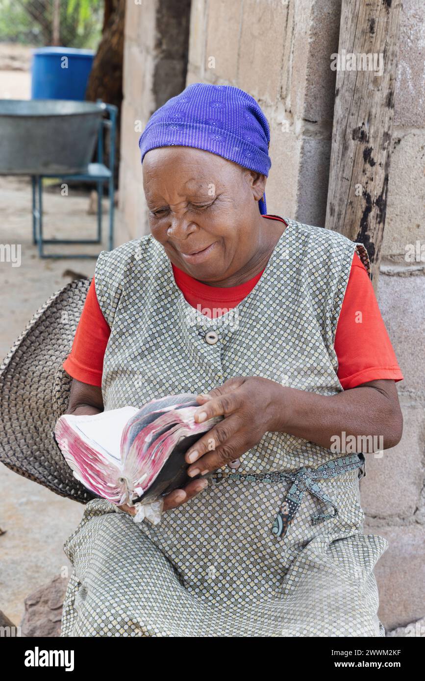 Heureux village religieux chrétienne africaine vieille femme lisant la Bible, assise dans sa cour, cabane en arrière-plan Banque D'Images