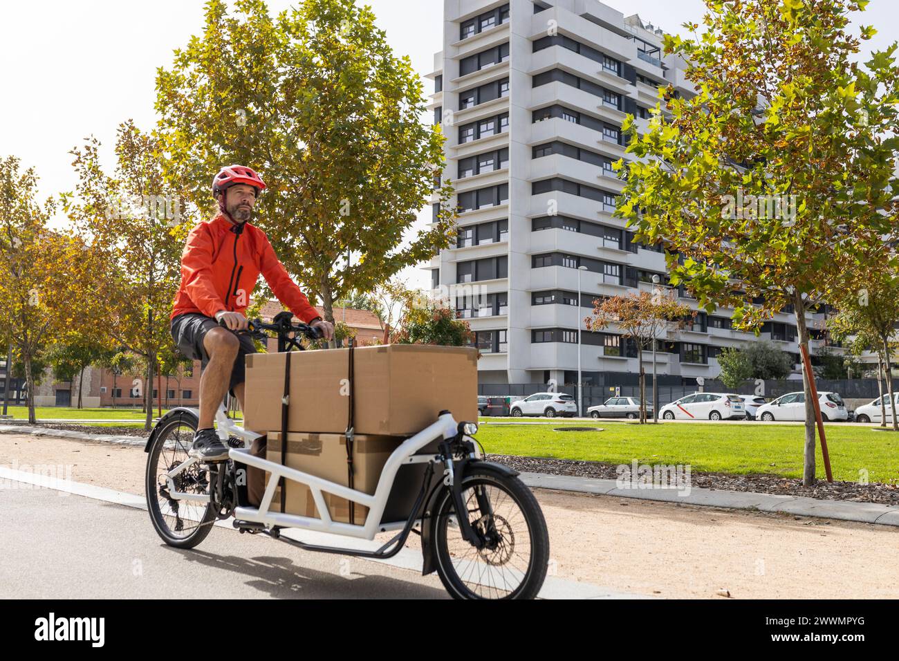 jeune courrier avec des vêtements rouges et casque vélo cargo, chevauchant le long de la piste cyclable de ville sur son chemin pour livrer un paquet. Banque D'Images