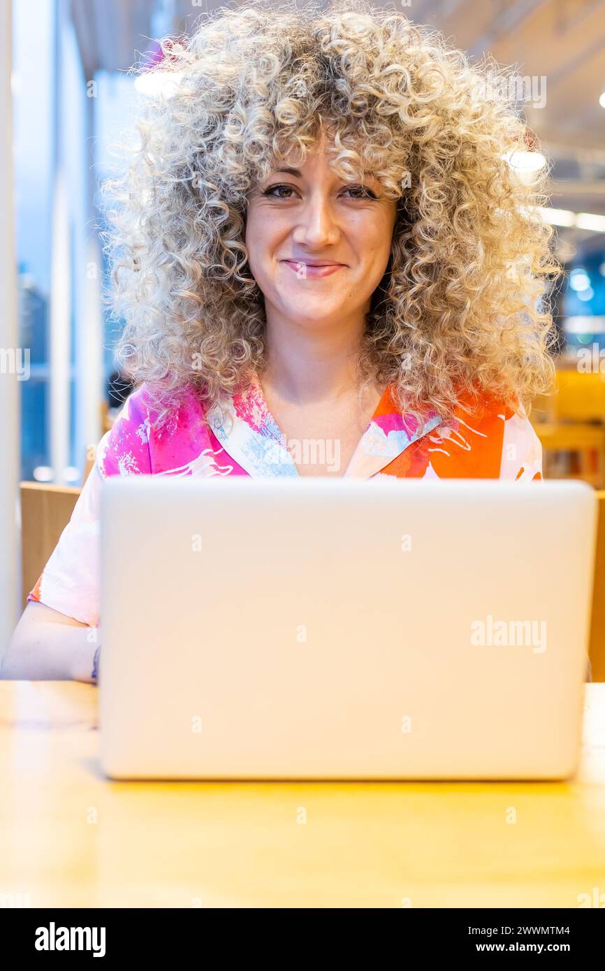 Portrait d'une jeune femme caucasienne souriante aux cheveux blonds bouclés, travaillant dans un café avec son ordinateur portable et son téléphone intelligent Banque D'Images