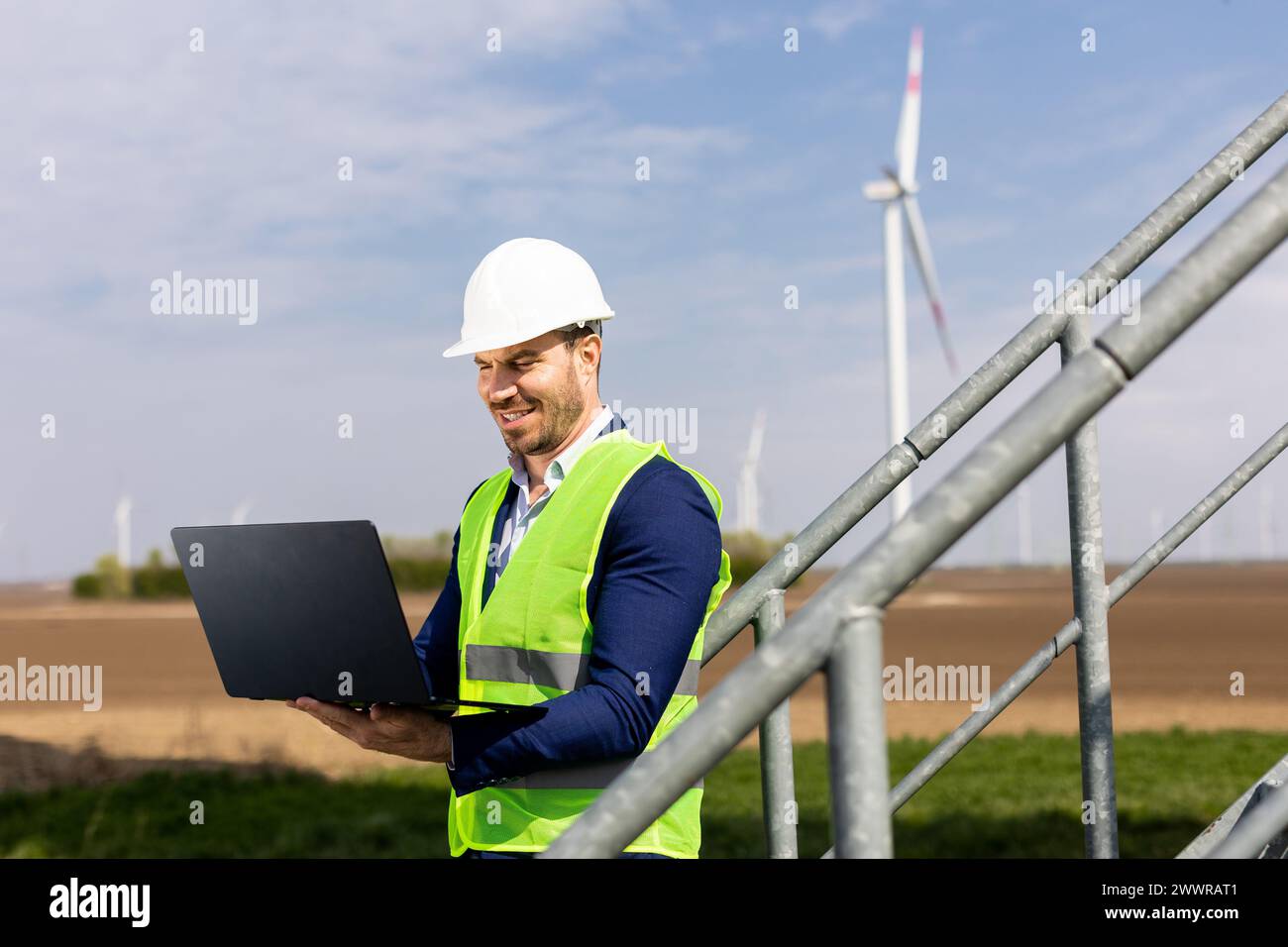 Un ingénieur spécialisé examine son ordinateur portable avec une éolienne en arrière-plan sous un ciel dégagé. Banque D'Images