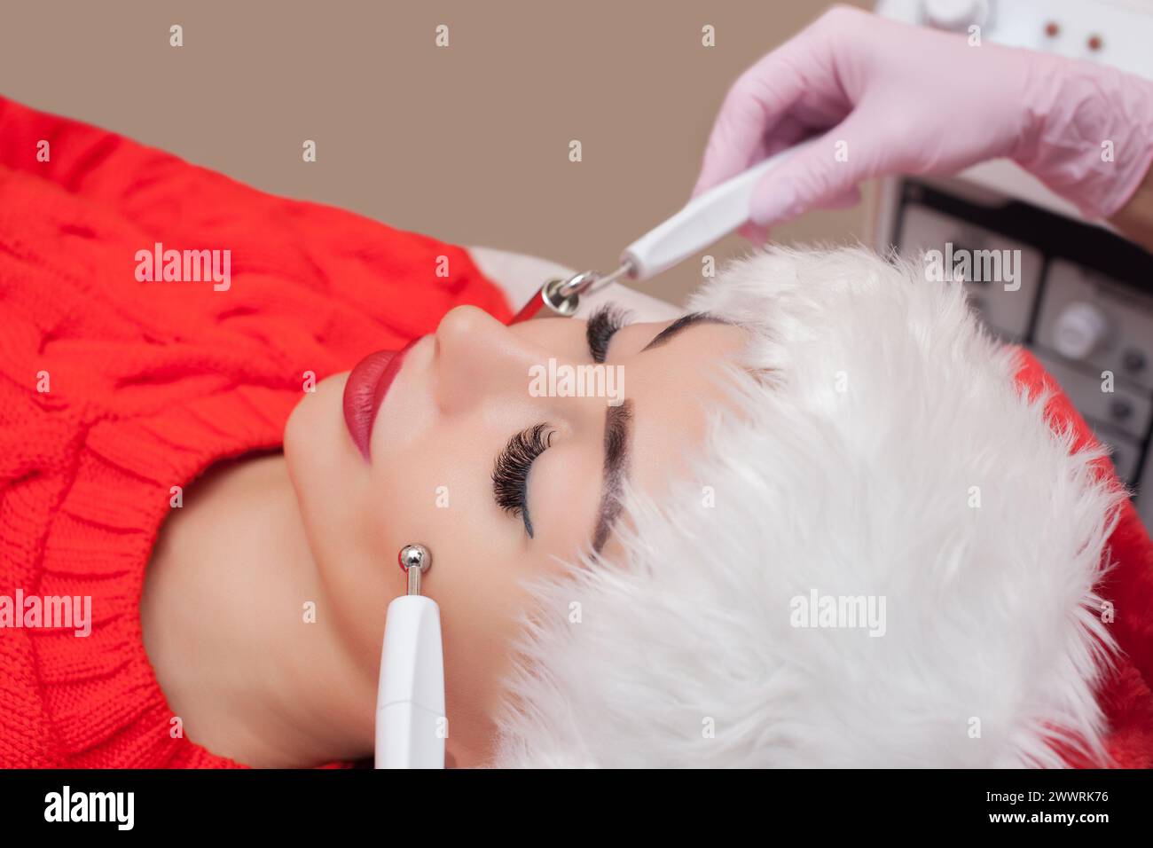 Le cosmétologue fait la procédure de thérapie par microcourant d'une belle jeune femme au chapeau du Père Noël dans un salon de beauté. Nouvel an et Cosmet Banque D'Images