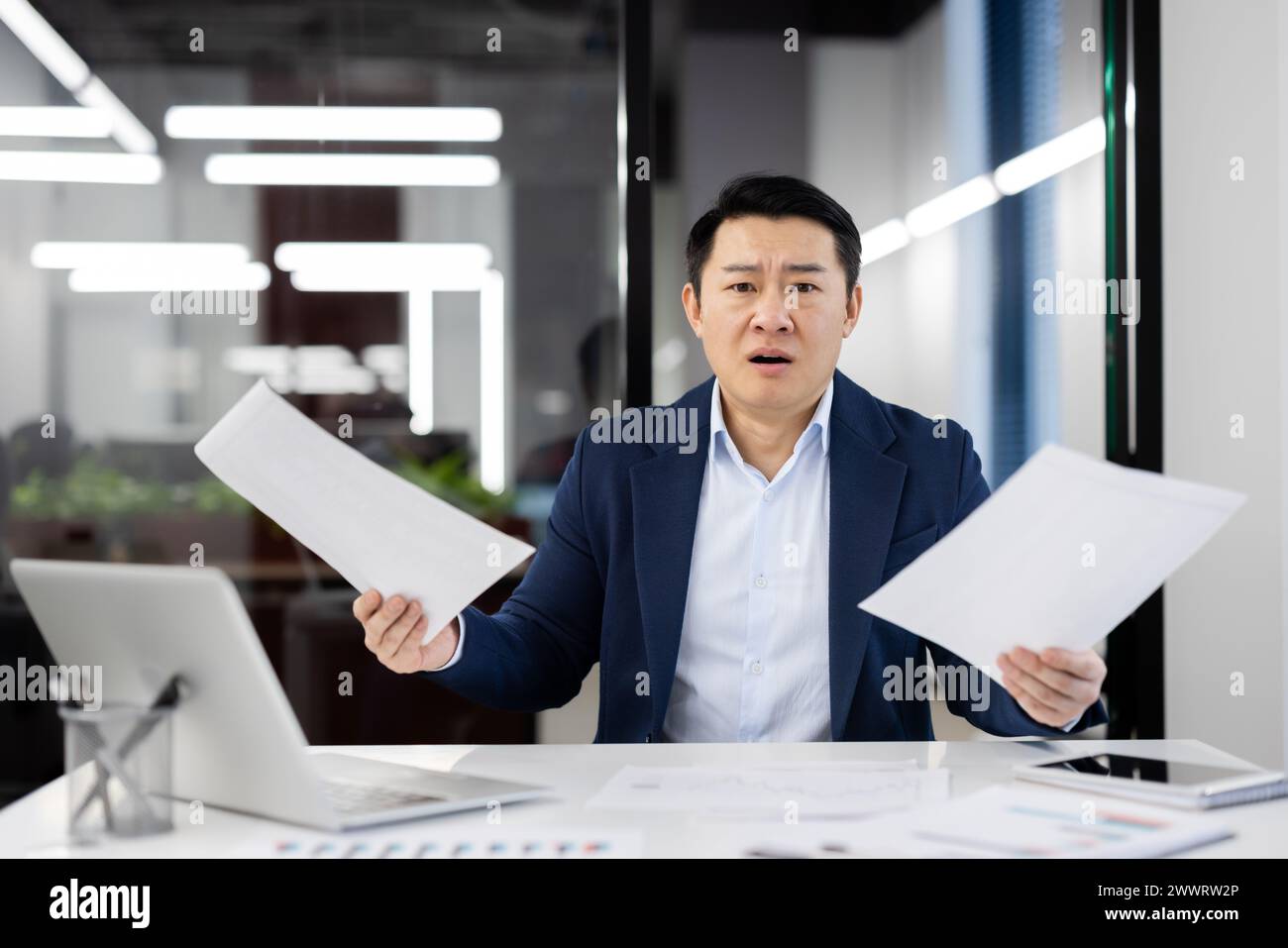 Un employé de bureau chinois choqué assis sur son lieu de travail personnel avec des documents en main. Homme perplexe utilisant un ordinateur portable moderne pour faire du travail en ligne à l'intérieur. Environnement de bureau. Banque D'Images