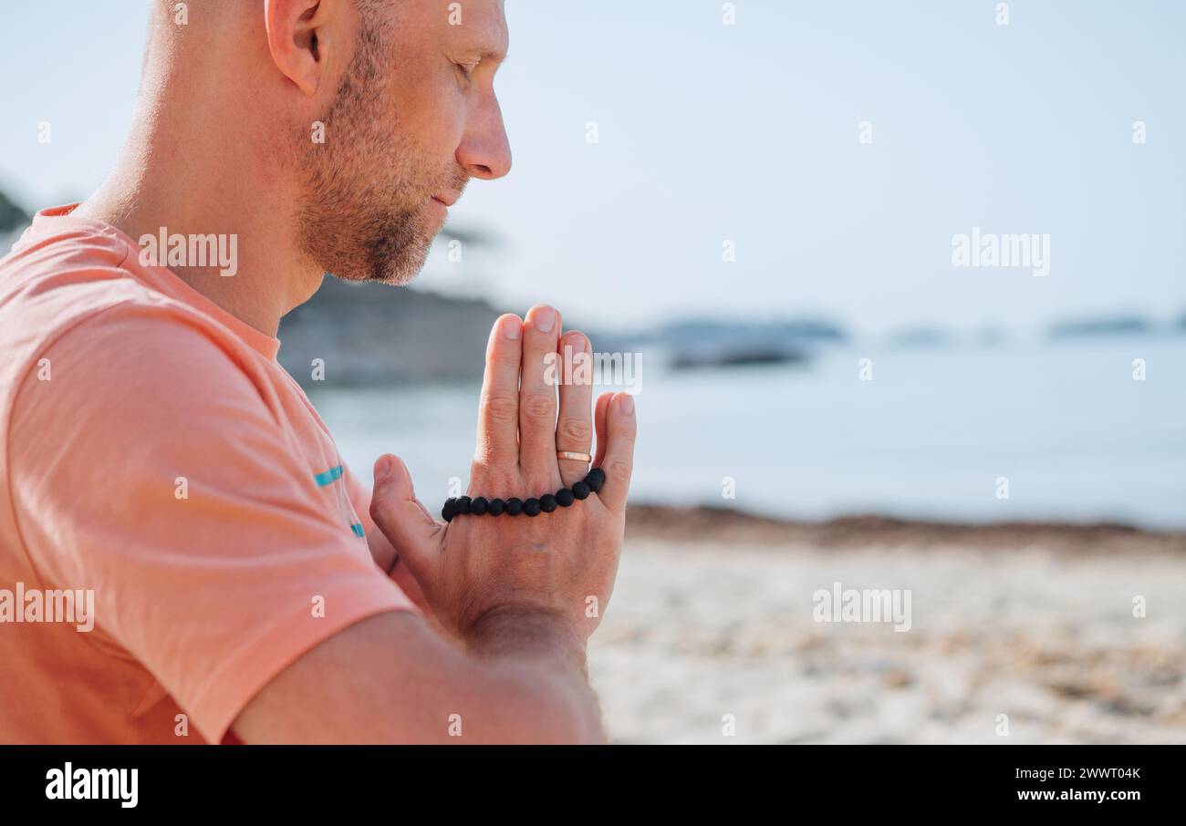 Homme calme assis avec les mains Namaste ensemble faisant des exercices de respiration profonde et méditant tôt le matin sur une plage de sable avec des vagues de mer calmes. Banque D'Images