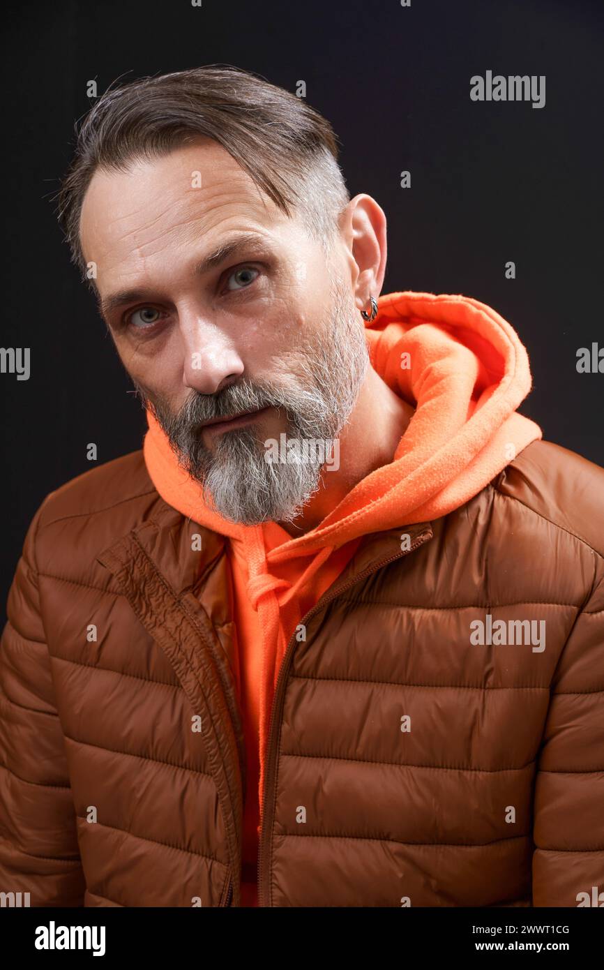 Un homme avec une barbe et des cheveux gris porte une veste marron et un sweat à capuche orange. Il regarde la caméra avec une expression sérieuse Banque D'Images