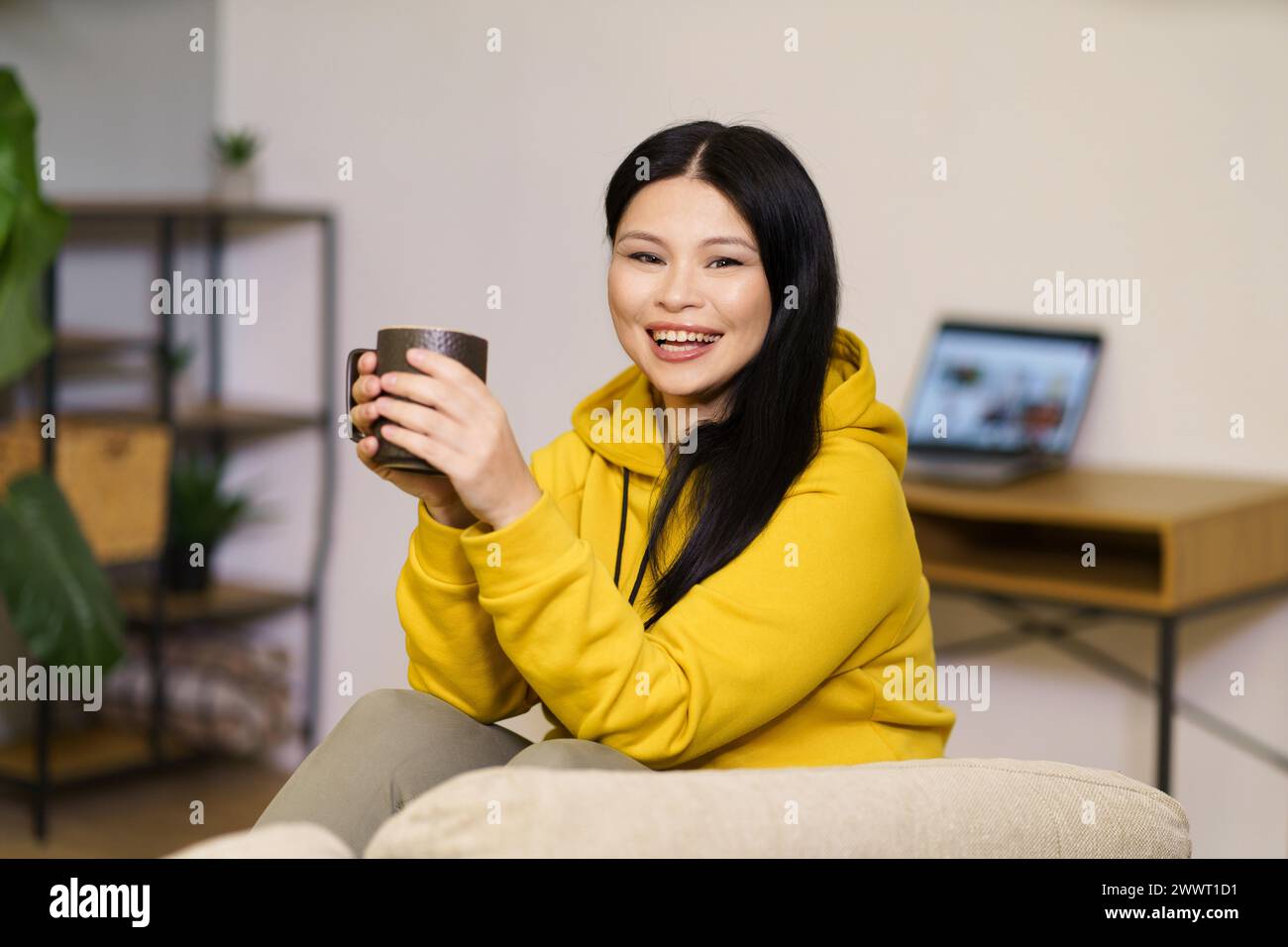 Une femme dans un sweat à capuche jaune sourit tout en tenant une tasse. La chambre a une atmosphère chaleureuse avec un canapé et une plante en pot Banque D'Images